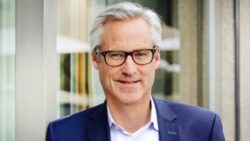 A smiling man with gray hair and glasses, wearing a dark blue blazer and a white shirt, stands in front of a modern glass facade; Copyright: VDMA/Anna McMaster
